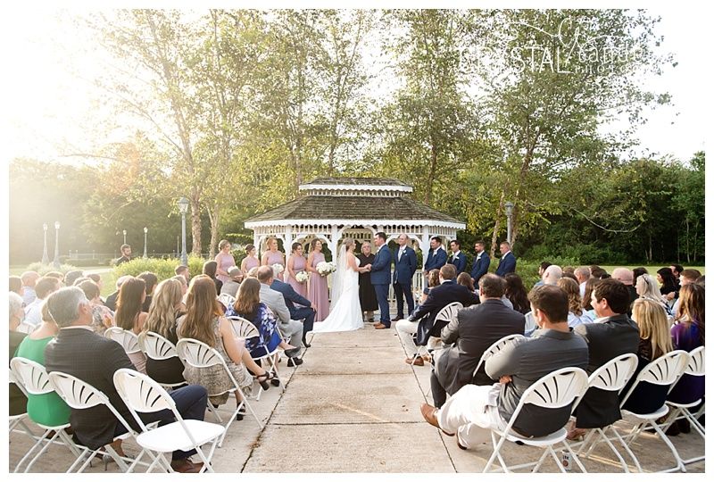 Outdoor wedding ceremony taking place at a white gazebo with guests seated in white chairs at sunset.