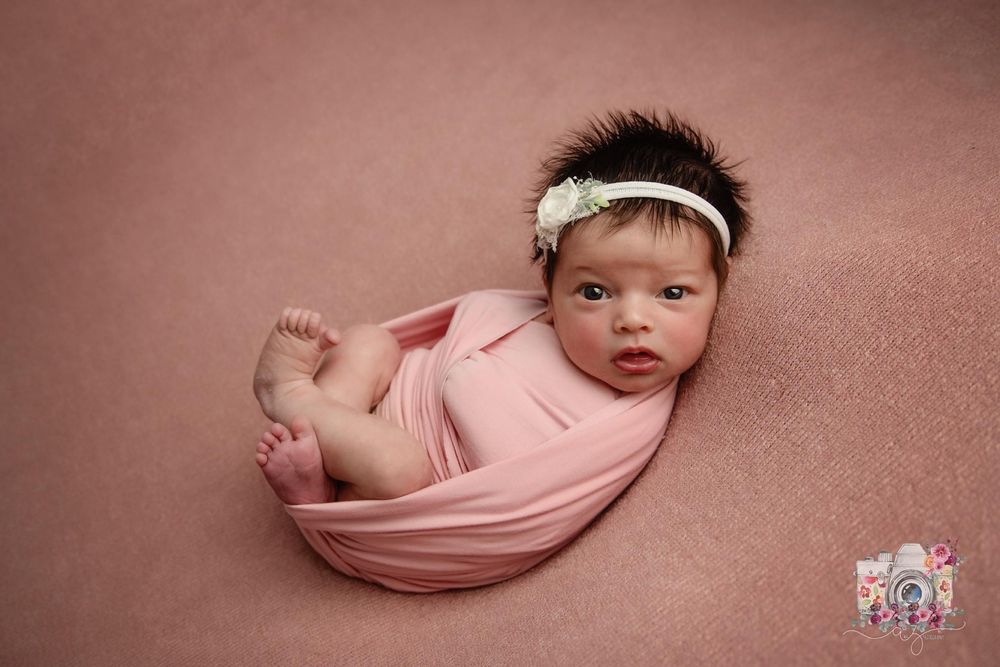 Alert newborn wrapped in pink fabric with white headband against rose backdrop.