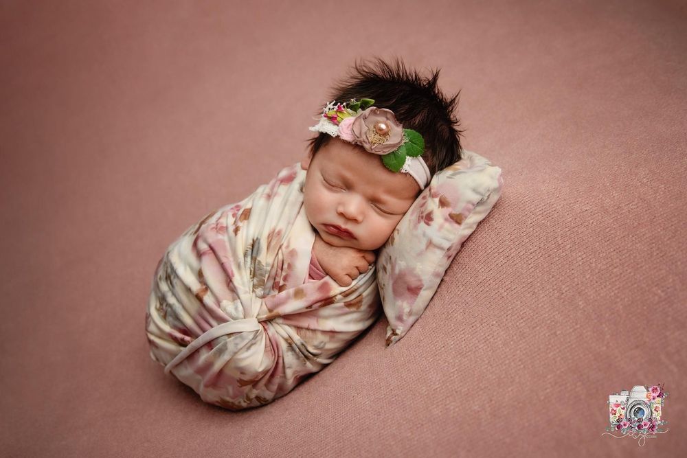 Sleeping newborn with dark hair wearing floral headband and patterned wrap on pink background.