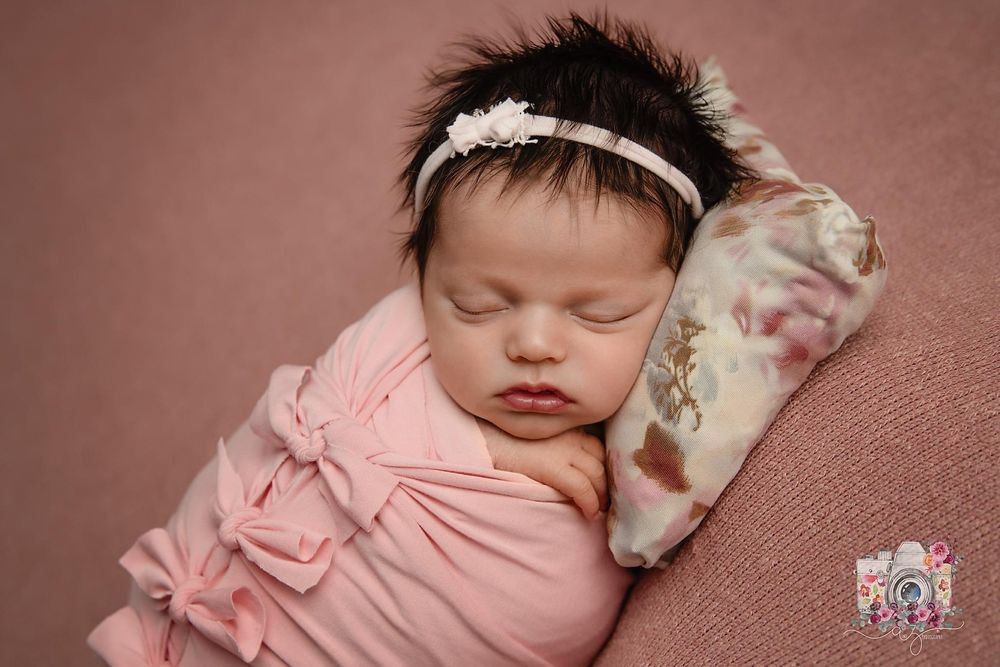 Newborn baby in pink wrap wearing lace headband against rose-colored backdrop.