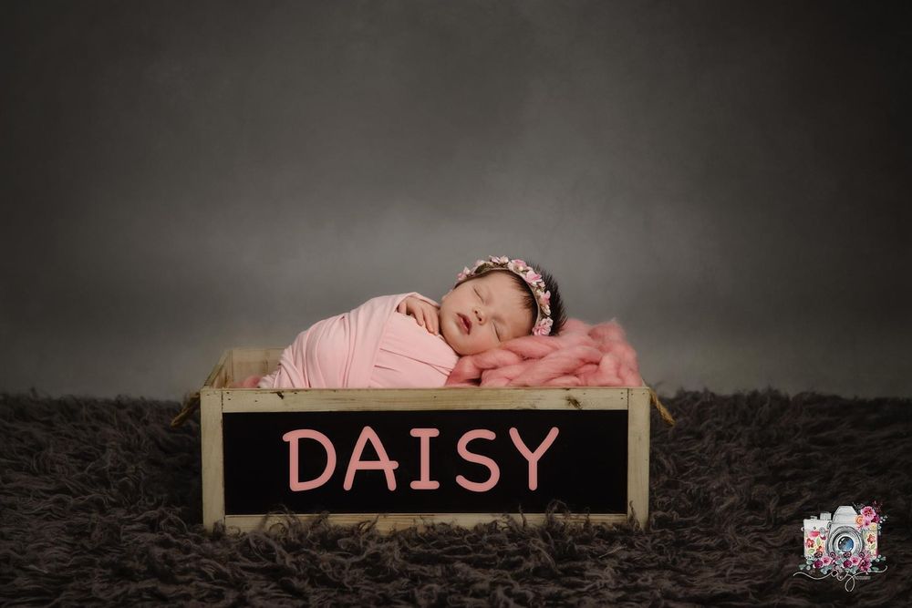 Newborn sleeping in pink wrap on wooden box with chalk sign reading Daisy against dark backdrop.