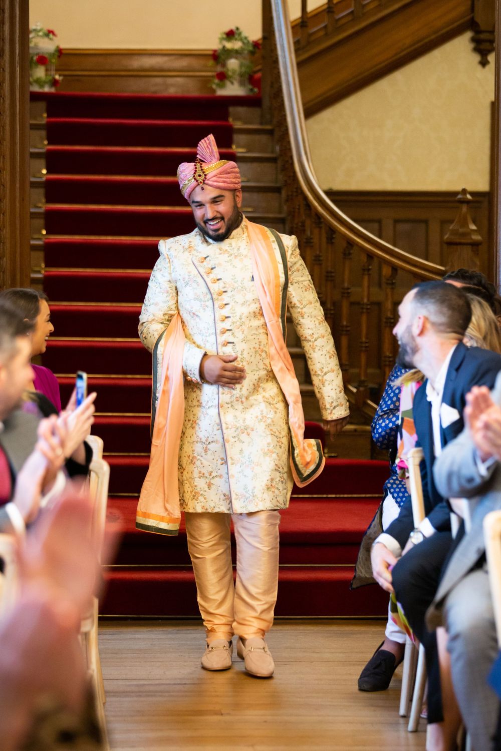 Groom in ornate gold and cream traditional outfit descends grand staircase while guests applaud.