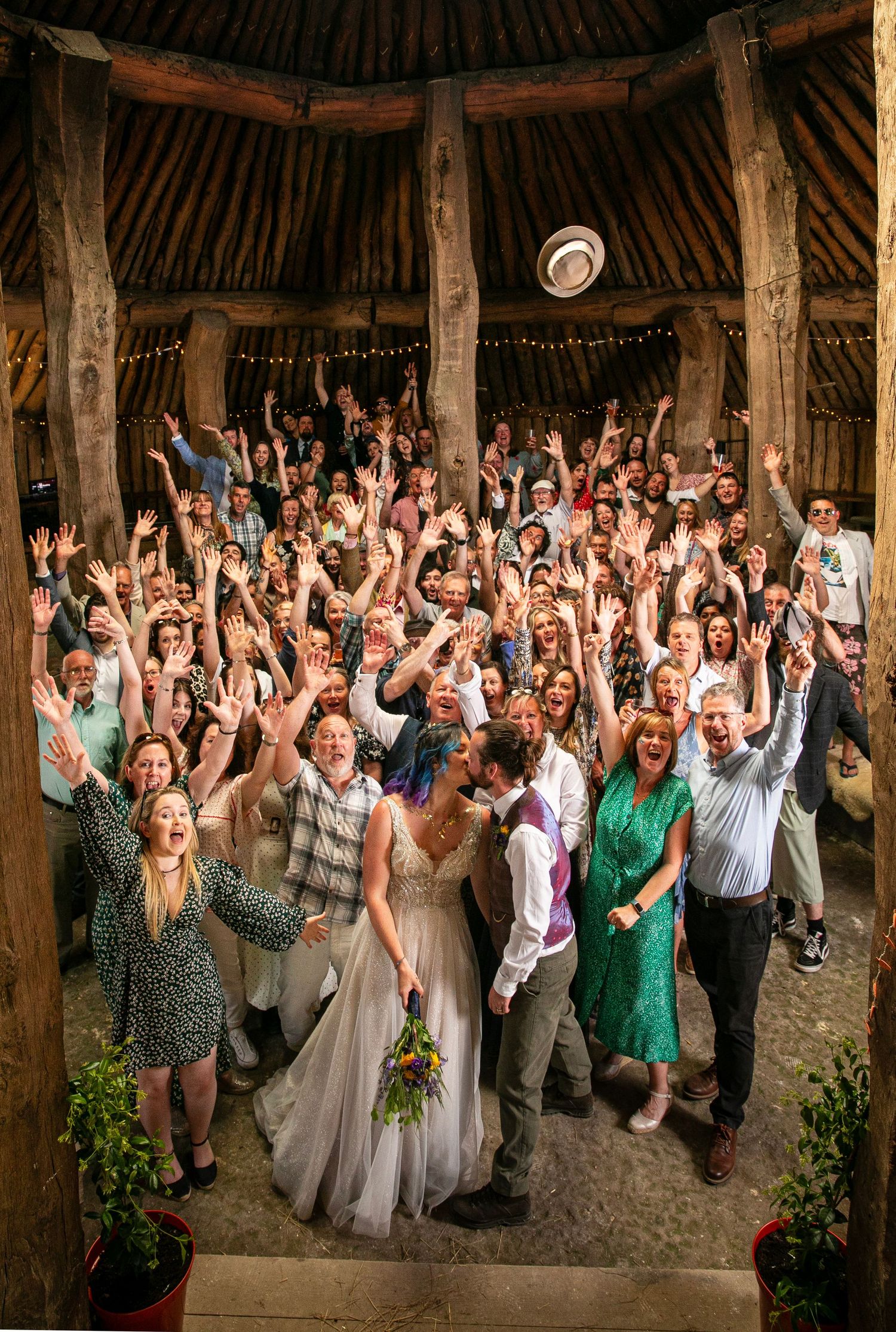Large group of wedding guests with raised arms celebrating in a rustic barn setting.