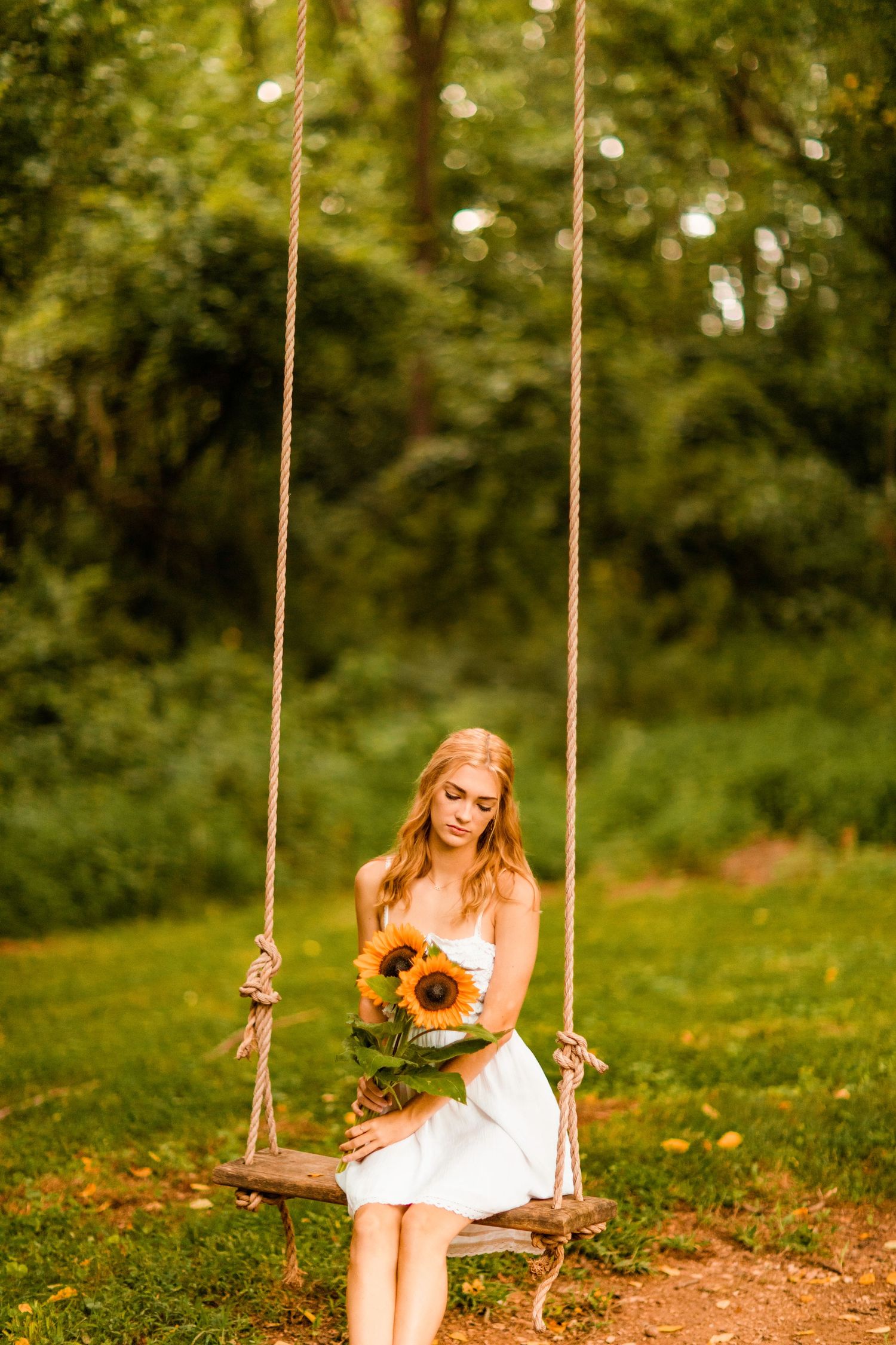 Young woman in white dress sits on rope swing holding sunflowers in lush green garden.