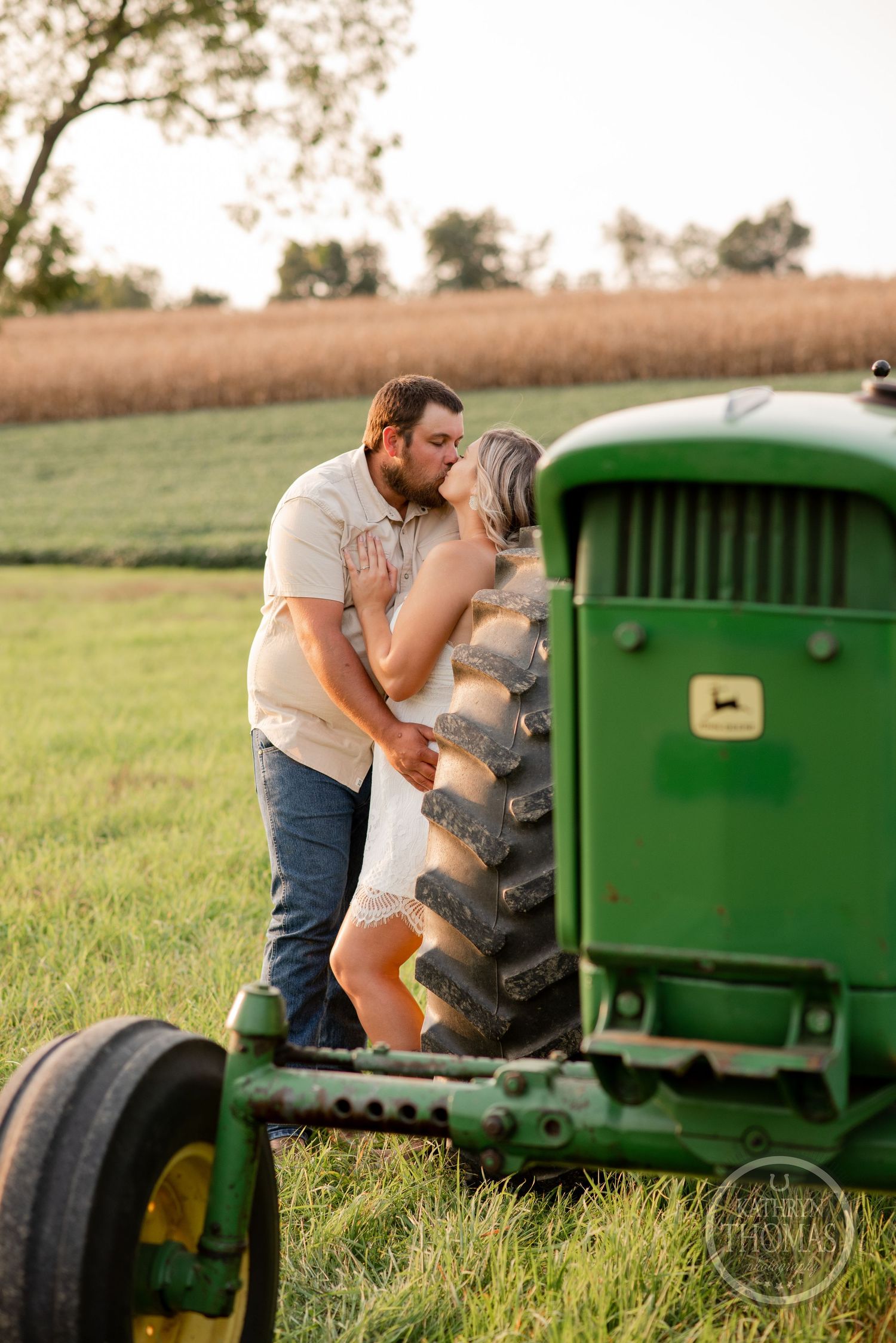 Romantic couple sharing an intimate moment by a vintage green John Deere tractor in a rural field at sunset.