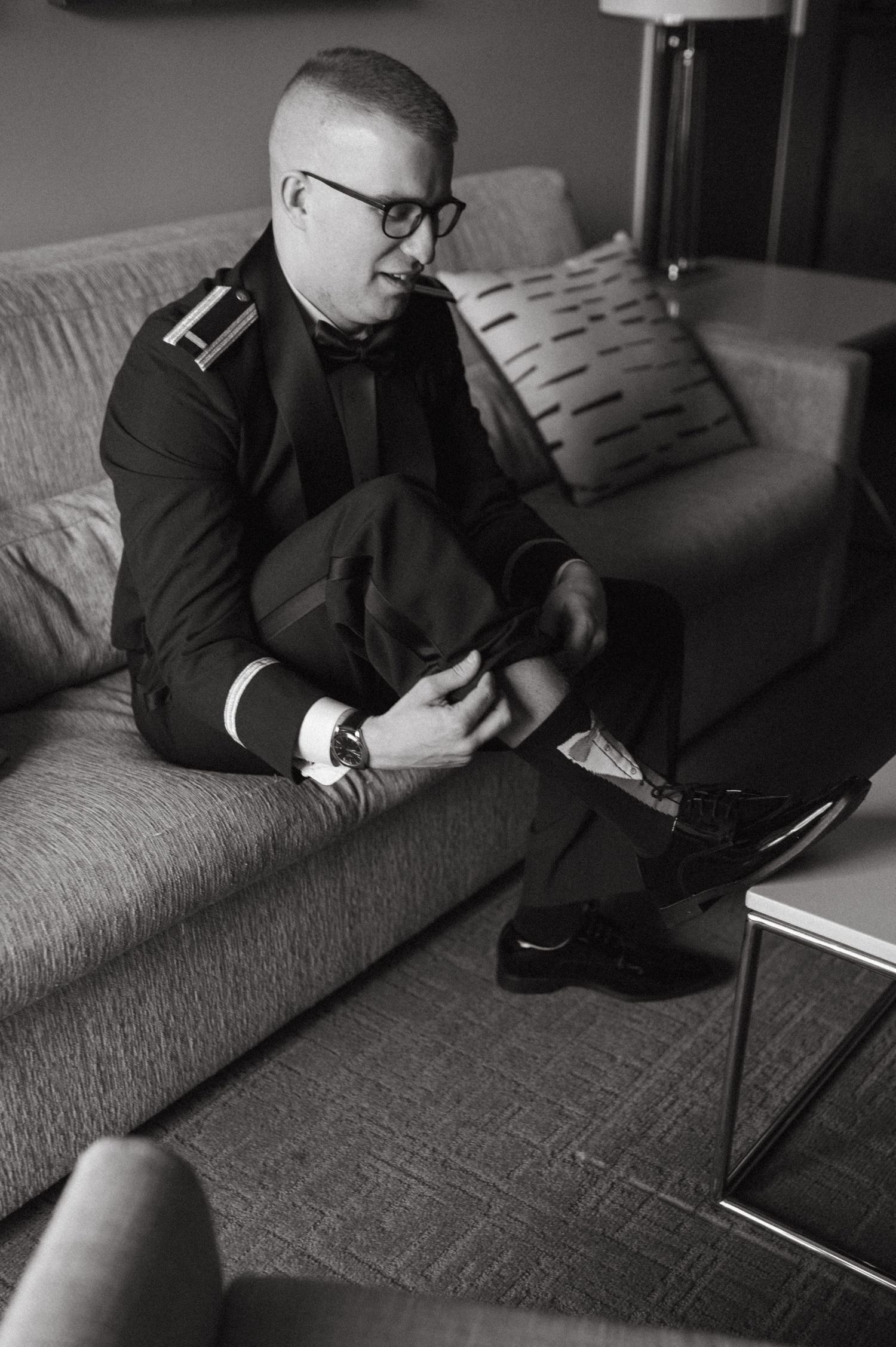 Black and white photo of a person in formal attire adjusting shoes while seated on a gray couch.