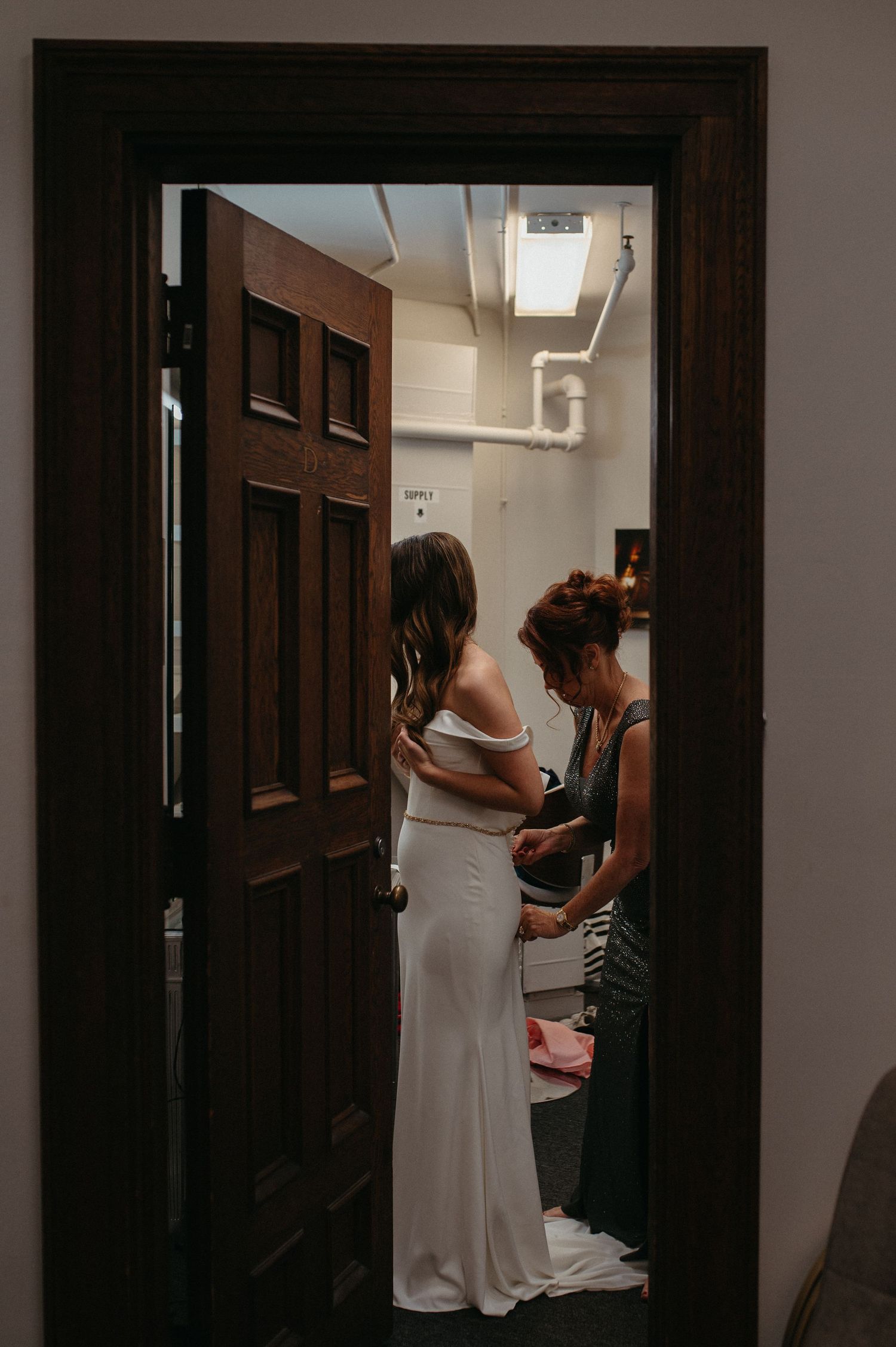 Wedding dress preparation scene through doorway showing final adjustments being made.