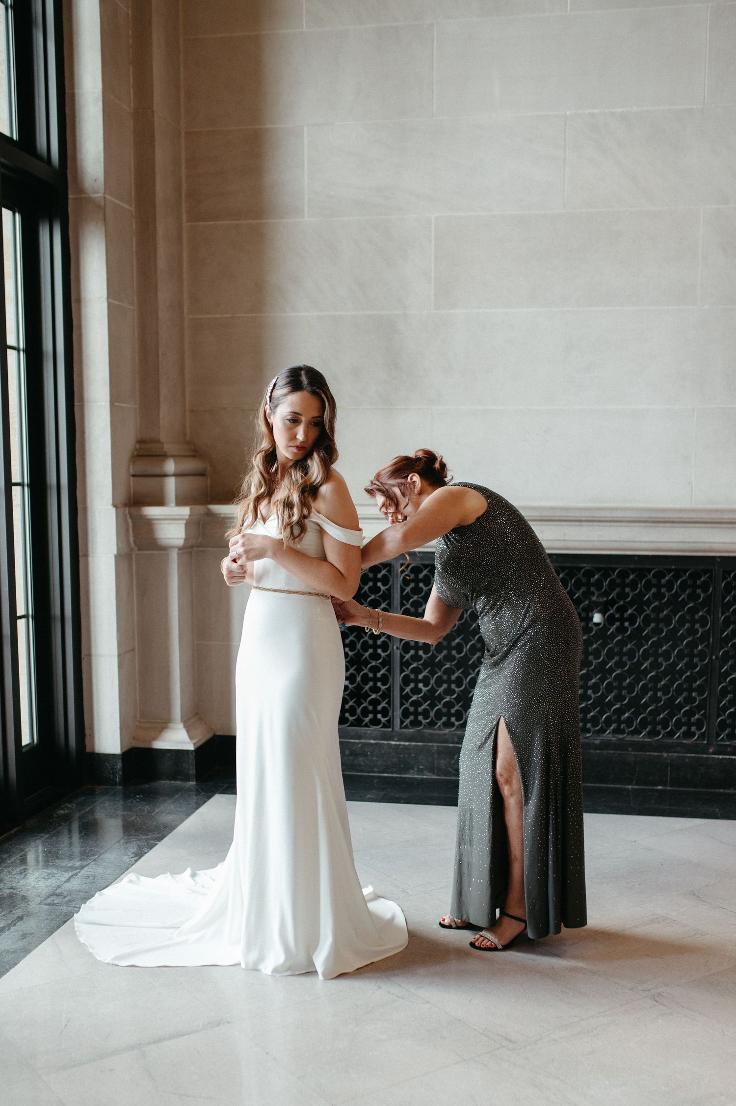 Wedding dress fitting moment as helper adjusts white gown in elegant interior.
