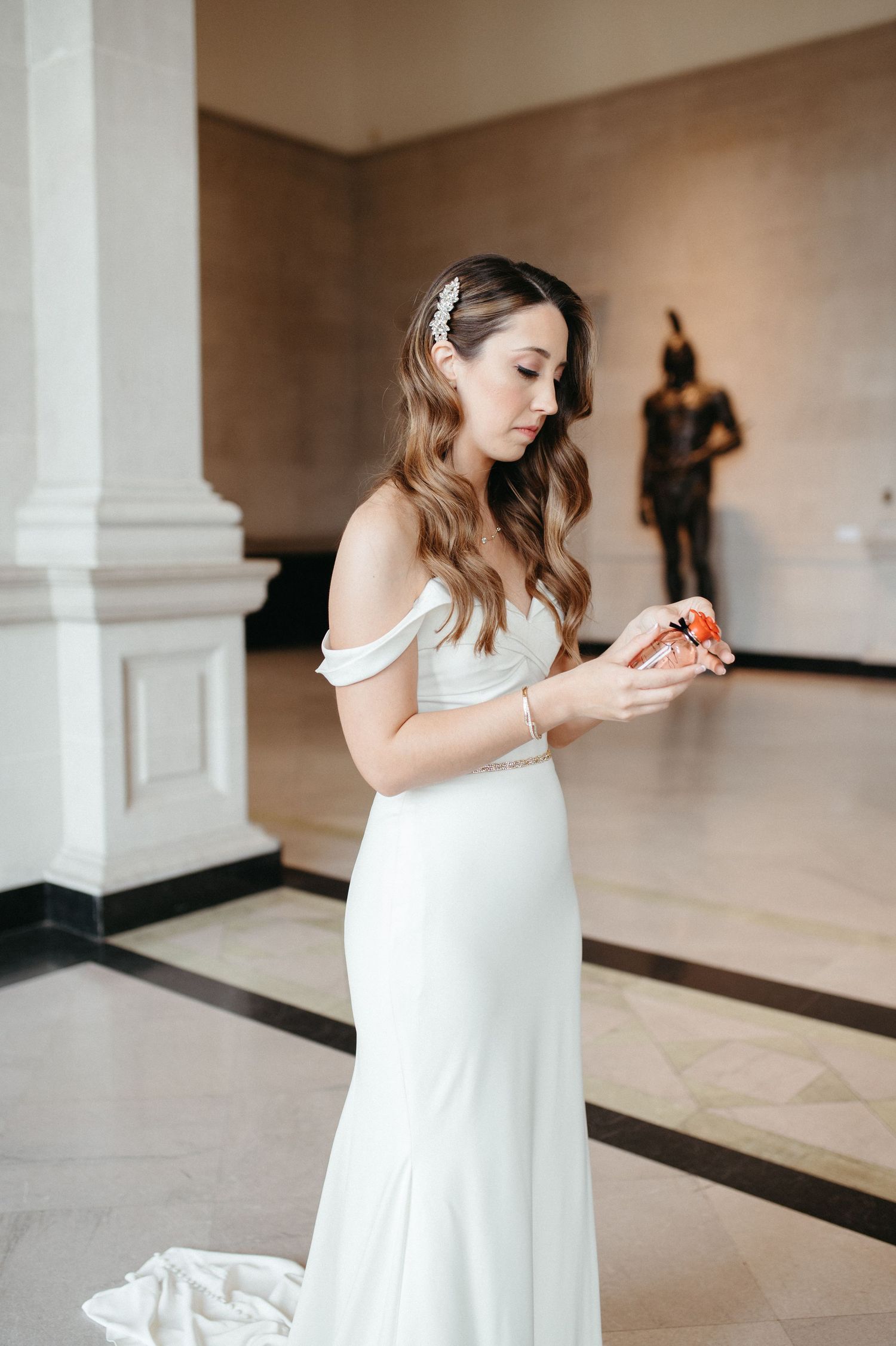 Elegant bride in off-shoulder white gown holding small object in marble hallway.