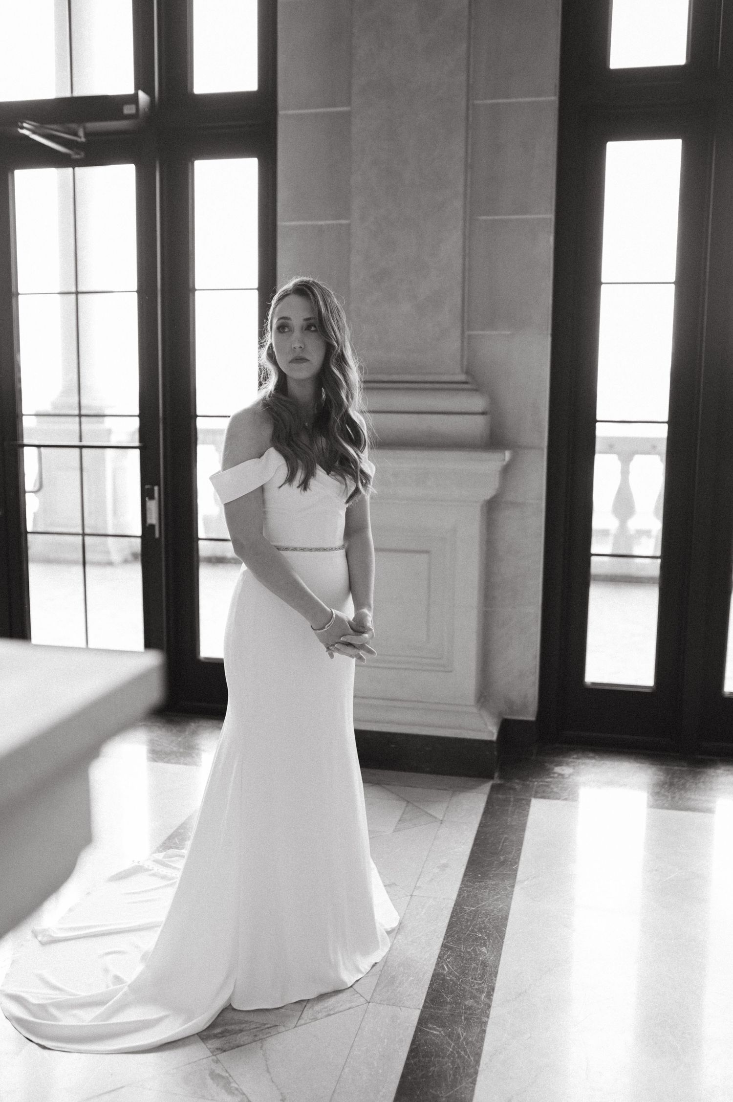 Black and white portrait of bride in flowing off-shoulder wedding gown by large windows.