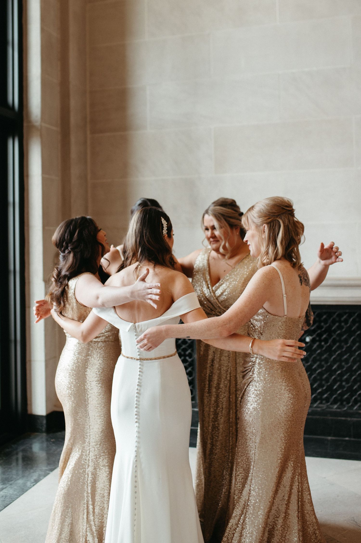 Bridesmaids in sparkly gold dresses embrace the bride in white gown during emotional pre-wedding moment.