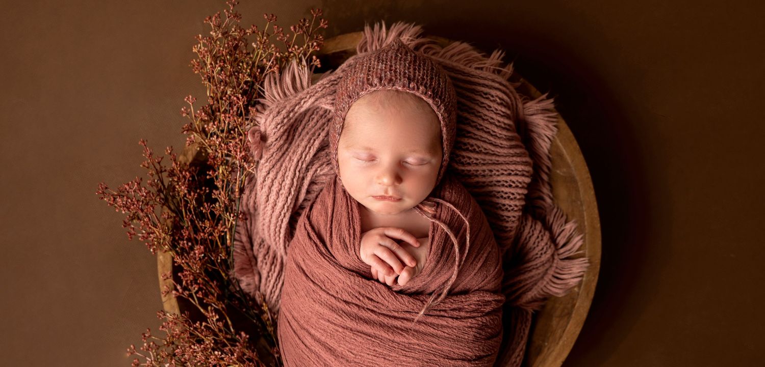 Newborn baby swaddled in dusty rose wrap and knit hat, sleeping peacefully in a wooden bowl with autumn botanicals.