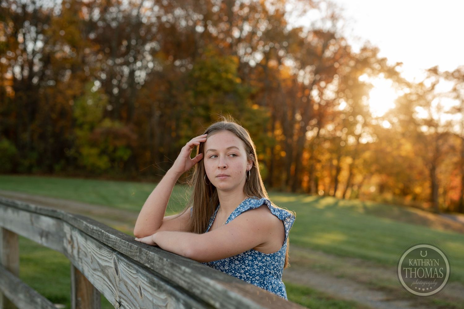 Someone in a blue dress leans on a wooden fence during golden hour with fall foliage in background.