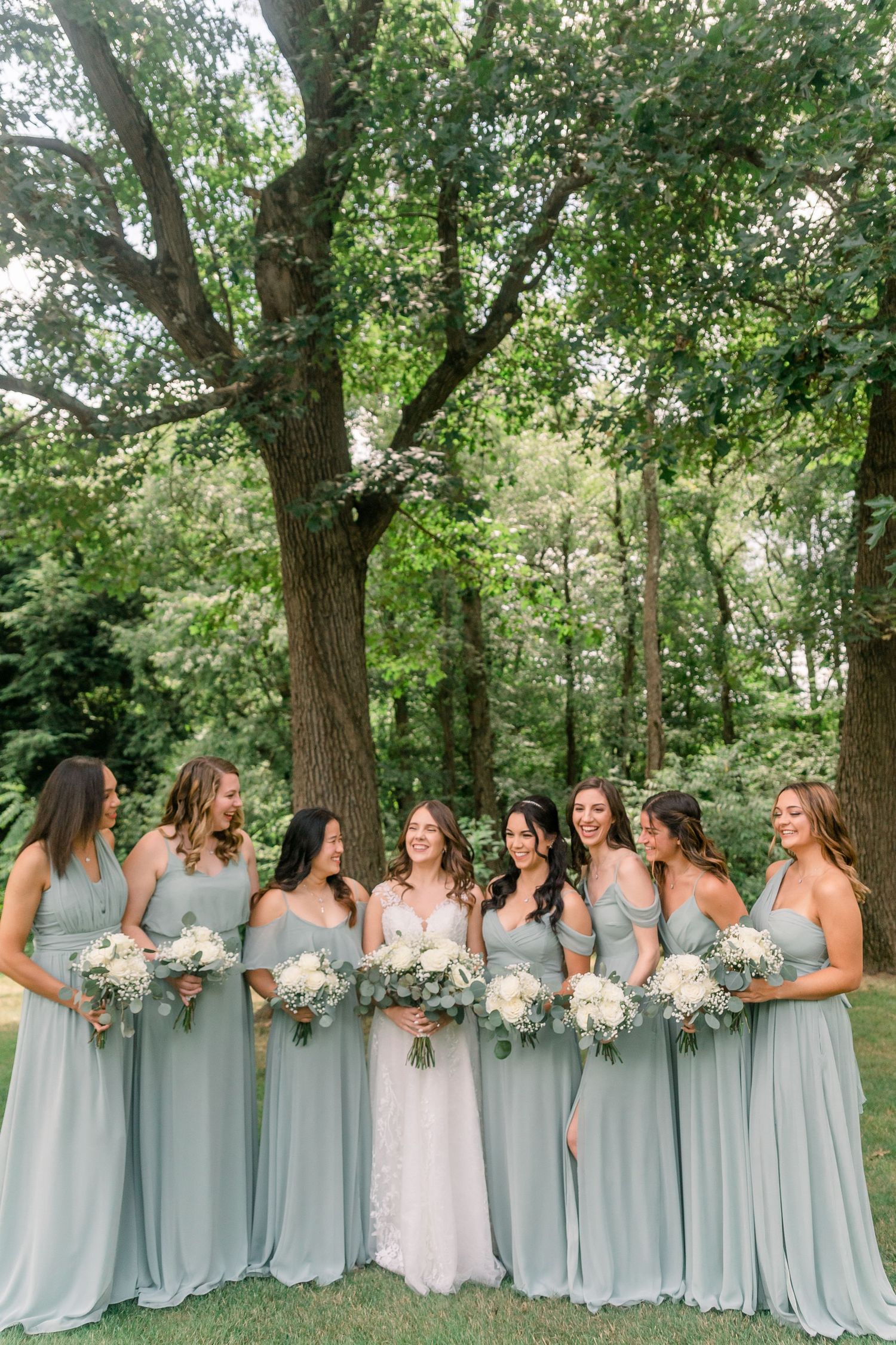 Large bridal party in light blue dresses poses together under green trees.
