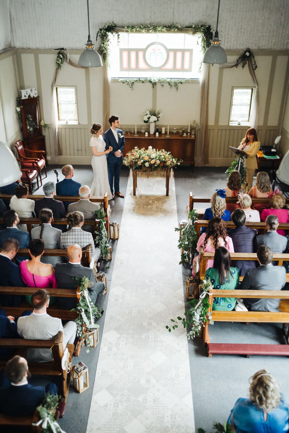 A wedding ceremony in a rustic chapel with wooden pews decorated with greenery and flowers.