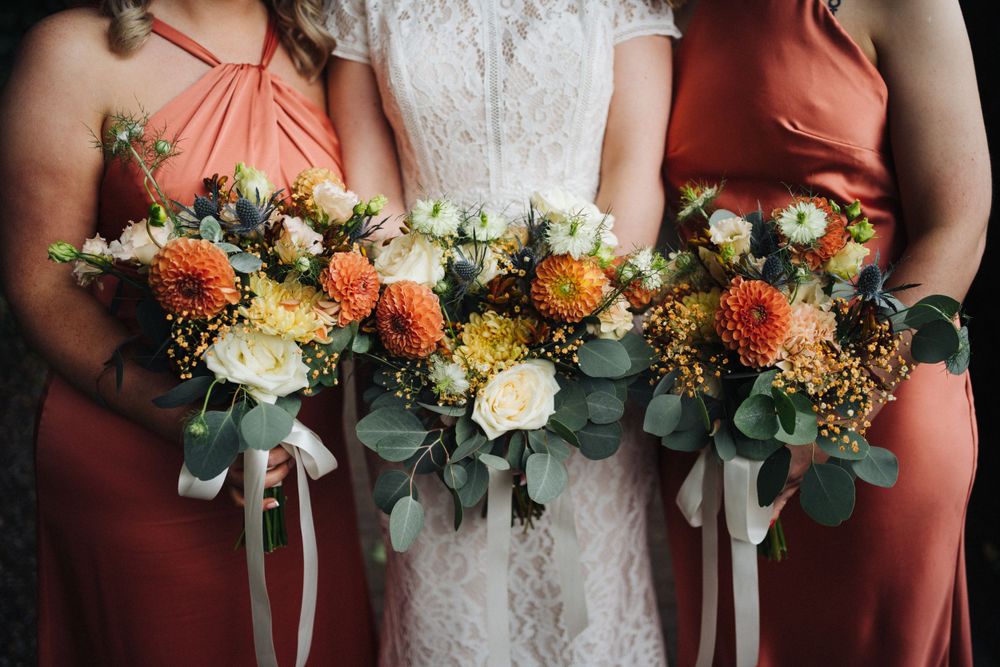 Beautiful autumn-colored wedding bouquets with orange dahlias and eucalyptus held against coral bridesmaid dresses.