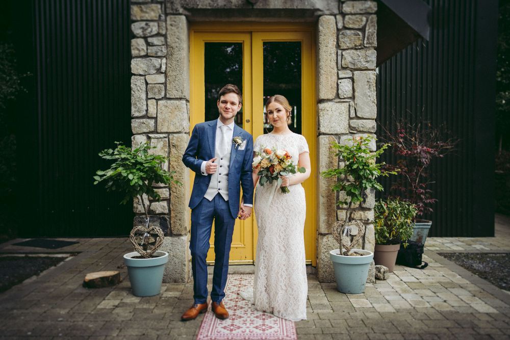Newlyweds pose in front of a charming stone building with yellow doors and potted plants.