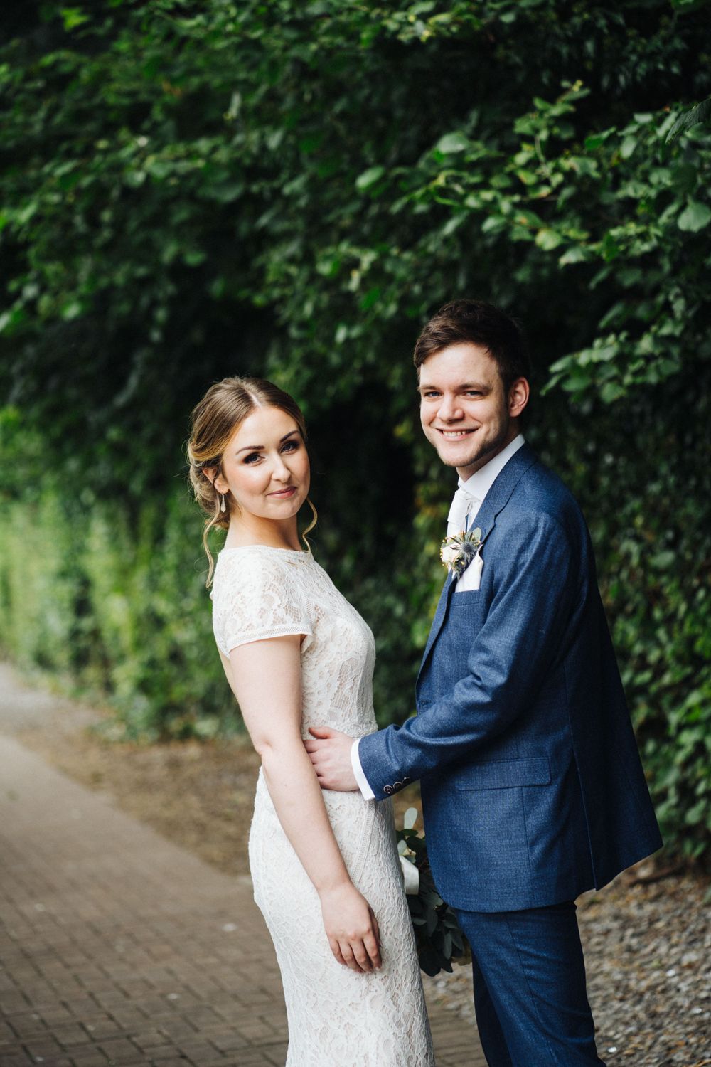 Elegant wedding couple poses on a brick pathway surrounded by lush greenery.