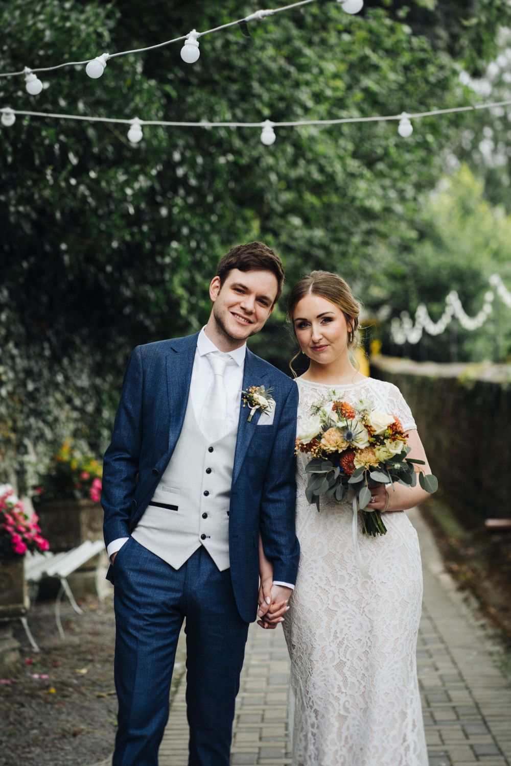 Wedding couple stands beneath string lights on garden pathway with stunning floral bouquet.