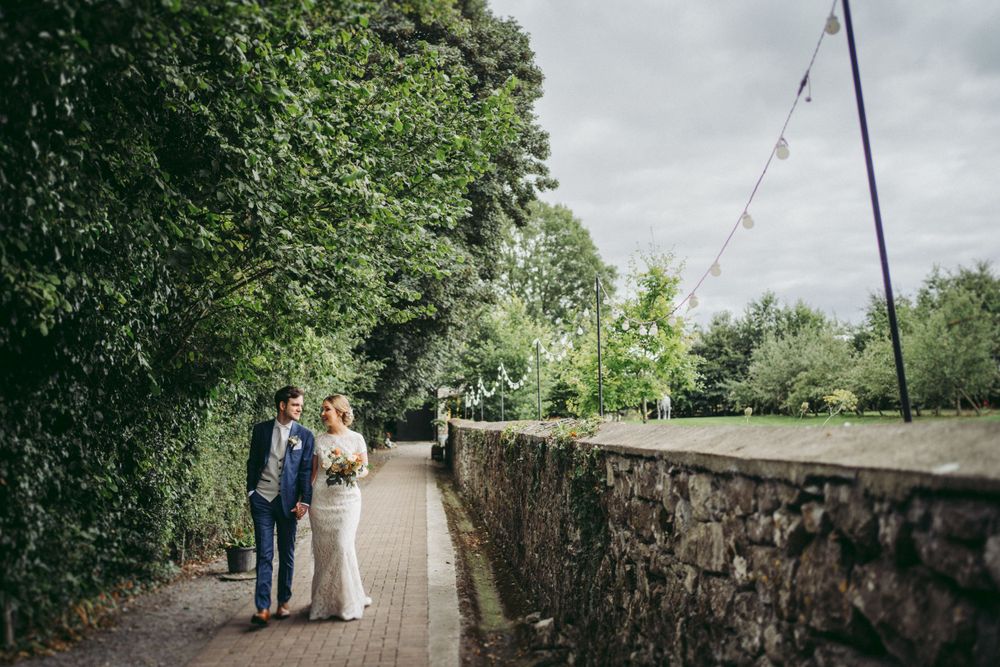 Couple walking along a stone wall path under cloudy skies with string lights overhead.