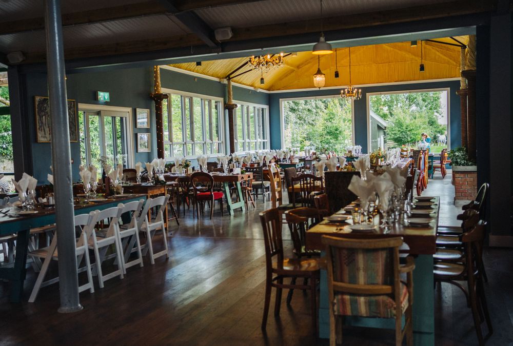 Spacious restaurant interior with wooden tables and chairs set against large windows overlooking greenery.