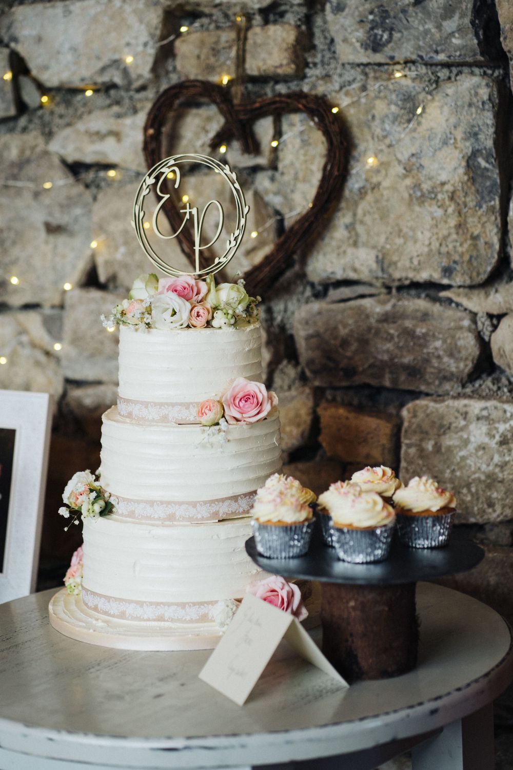 Three-tiered white wedding cake decorated with pink roses and gold topper beside cupcakes on stone backdrop.