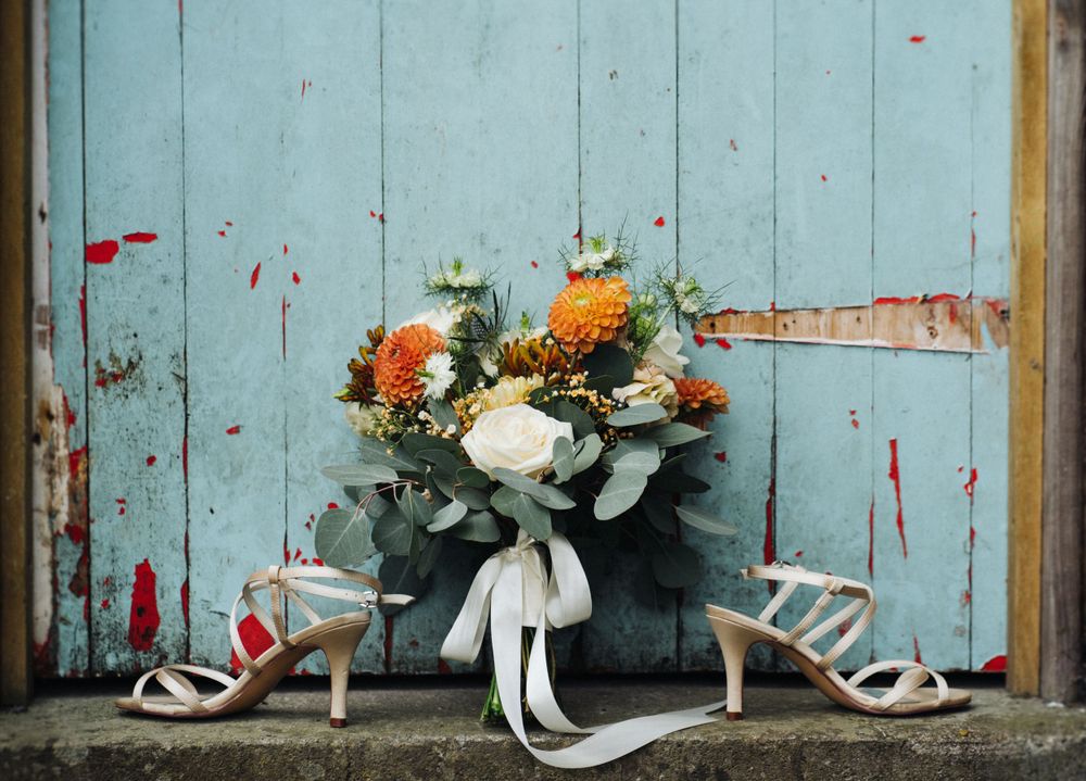 A bridal bouquet with orange and white flowers rests alongside elegant white heels against a weathered blue door.