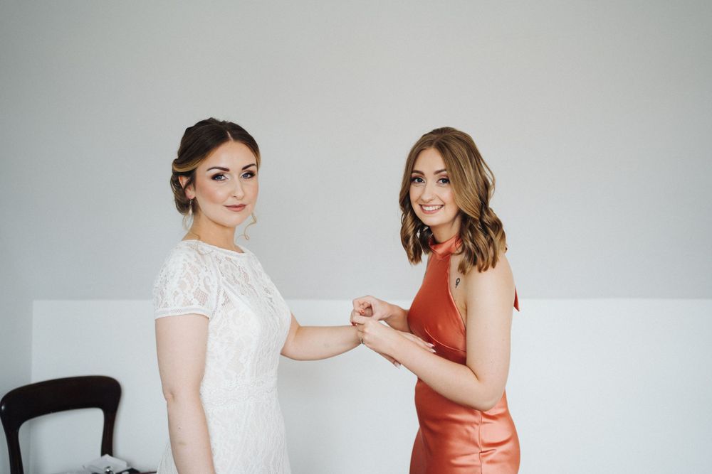 Two people pose together, one wearing a white lace dress and another in a coral-colored dress.