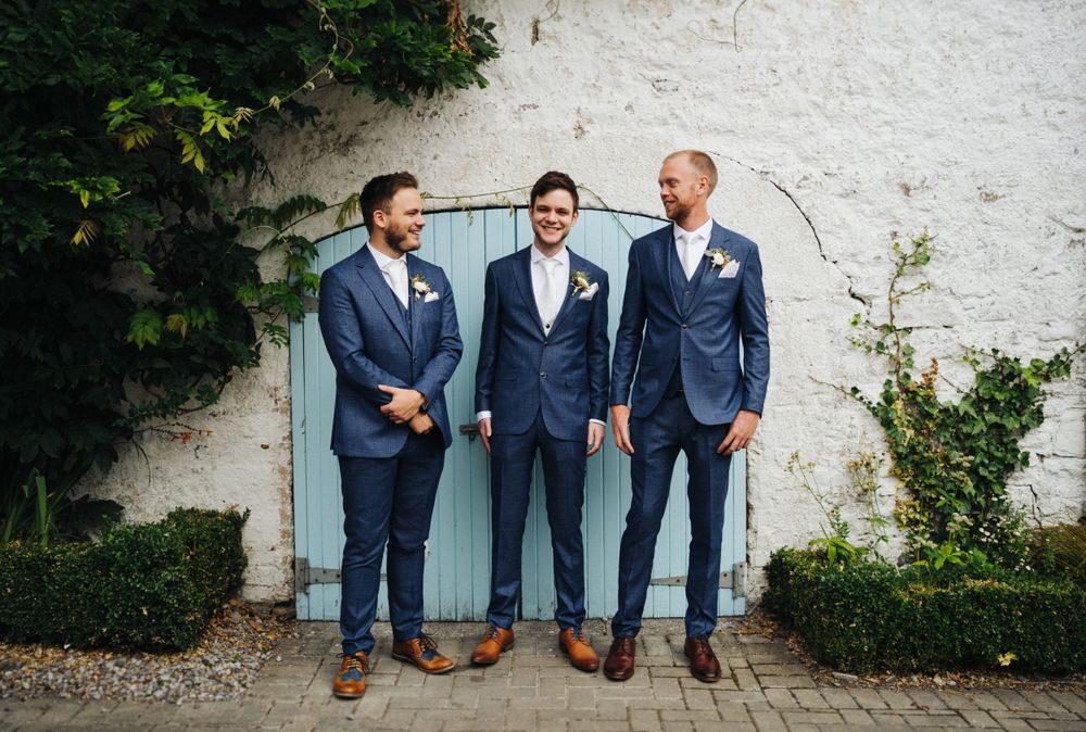 Three groomsmen in navy suits stand against a white wall with a blue door.