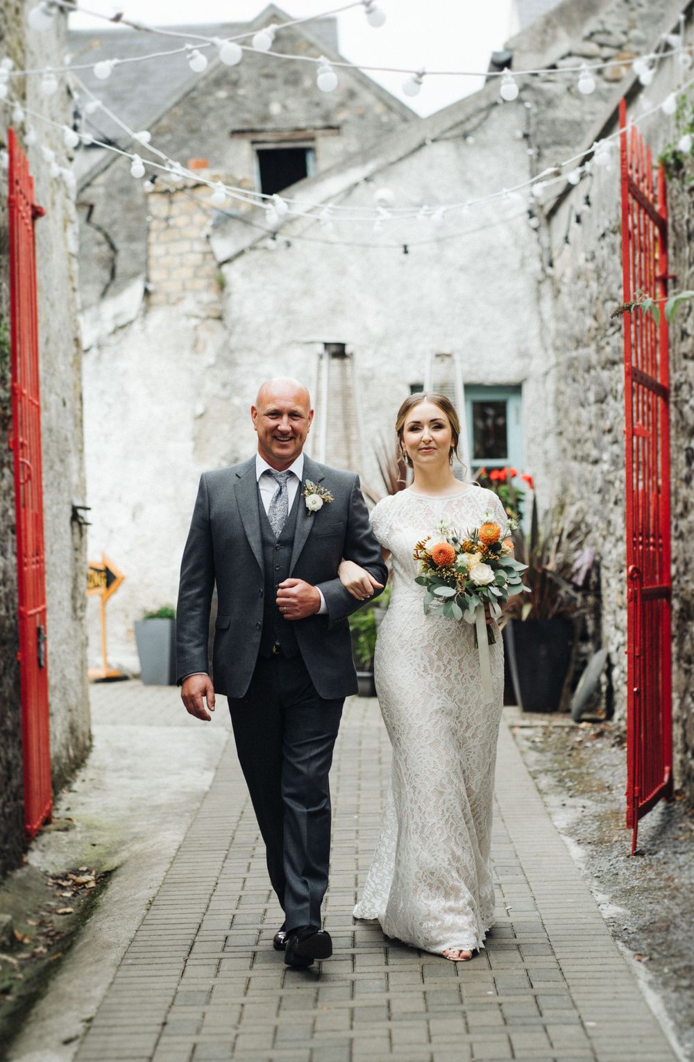 A couple walks down a quaint alleyway with white walls and red gates on their wedding day.
