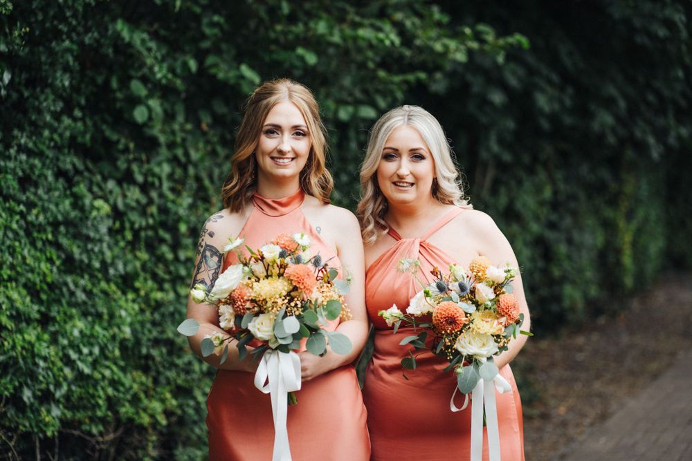 Two people in coral dresses hold matching floral bouquets against a green hedge background.