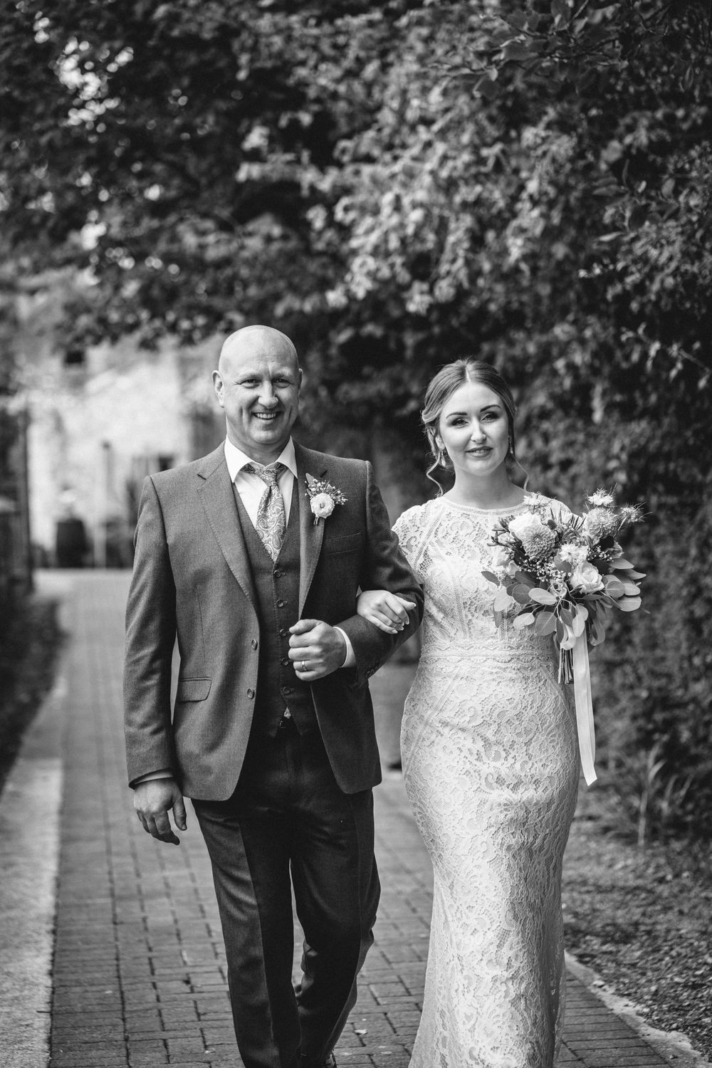 A couple walks together on a brick pathway surrounded by lush greenery on their wedding day.