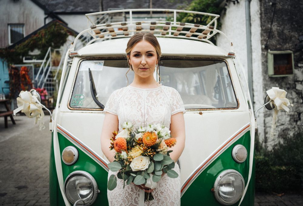 A bride in a lace dress poses in front of a classic green and white VW bus.