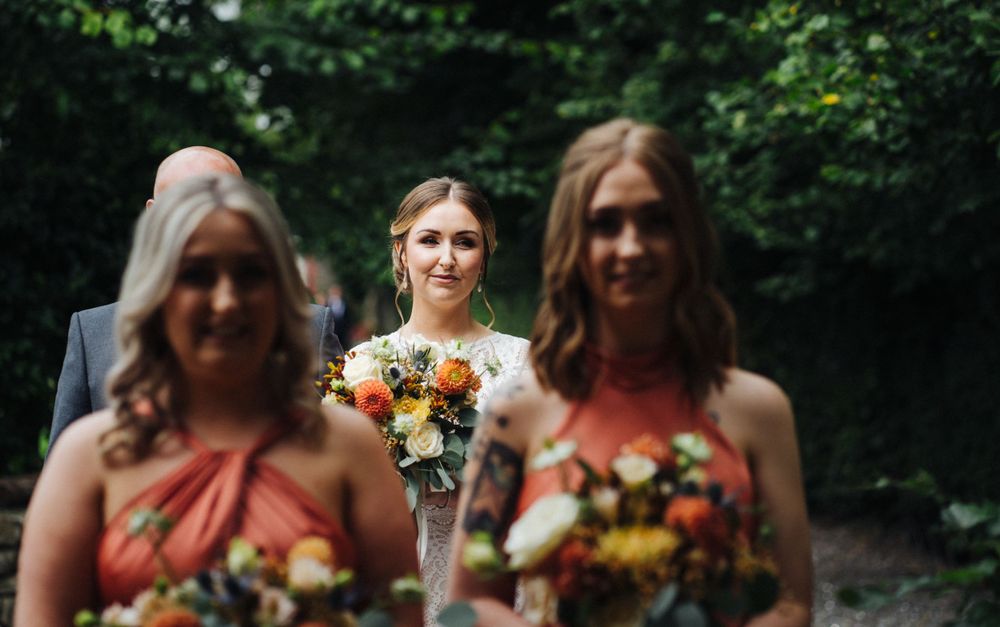 A wedding party walks down a tree-lined pathway with bridesmaids in coral-colored dresses.