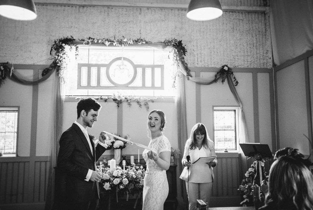 A black and white wedding ceremony photograph under decorative lighting with floral arrangements.