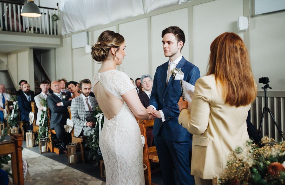 A wedding ceremony takes place in a bright indoor venue with guests seated in rows.