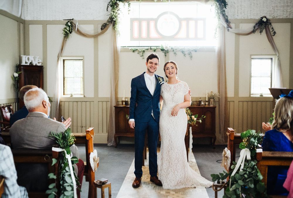 A wedding couple stands together in front of a large window in a rustic venue decorated with greenery.