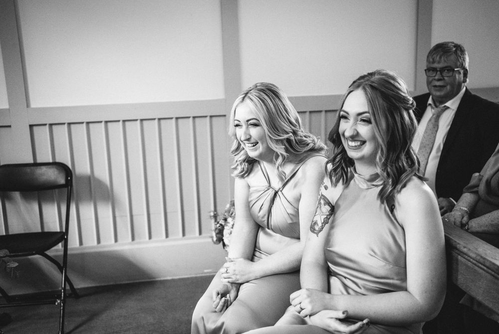Wedding guests share a joyful moment while seated on wooden benches in a bright venue.