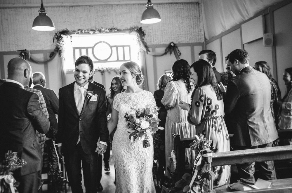 A black and white photo of a wedding couple walking through their venue surrounded by guests.