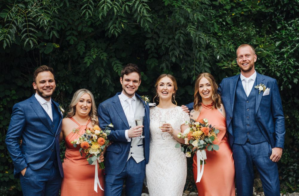 Bridal party in navy suits and coral dresses pose together outdoors with colorful bouquets.