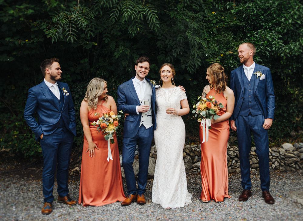 Wedding group stands together on gravel path wearing coral dresses and navy suits.