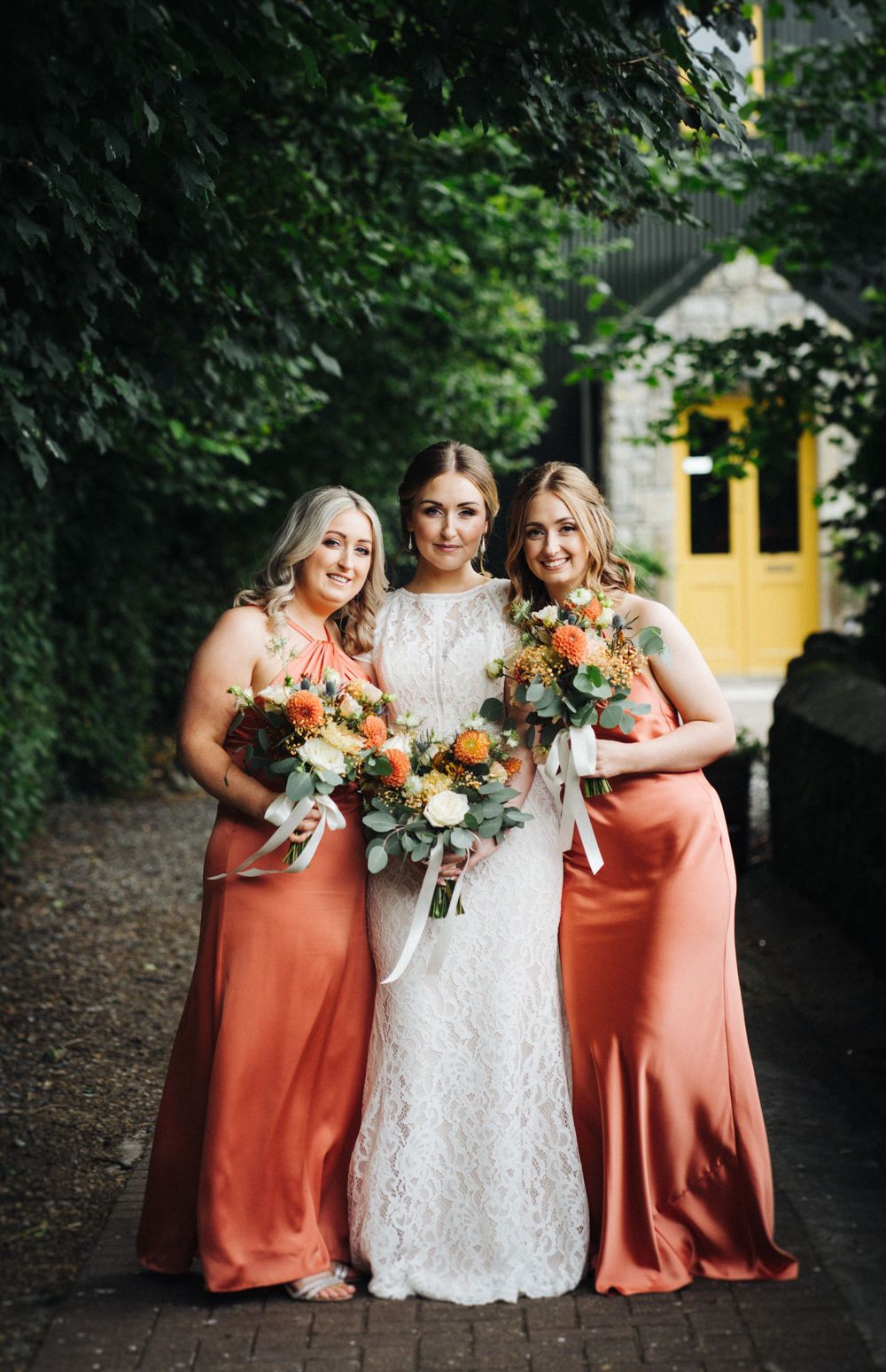 Three women pose in white lace and coral dresses holding autumn-toned bouquets.
