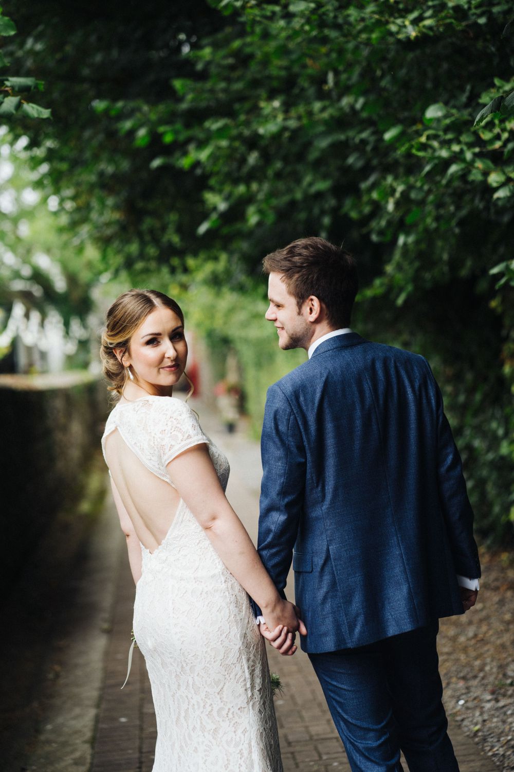 Couple walks hand in hand on garden path with bride in lace dress looking back at camera.