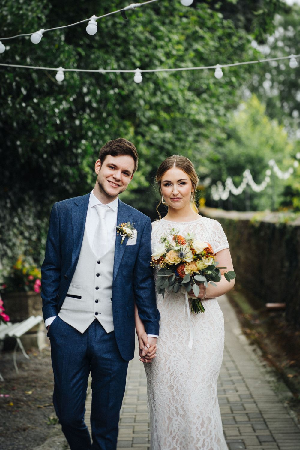 Newlyweds pose under string lights on garden path with bridal bouquet.