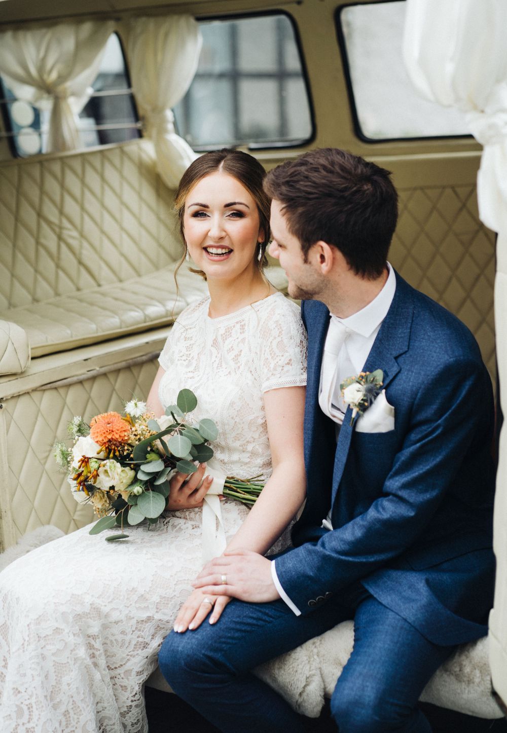 Couple sits together in vintage vehicle with quilted interior holding wedding bouquet.