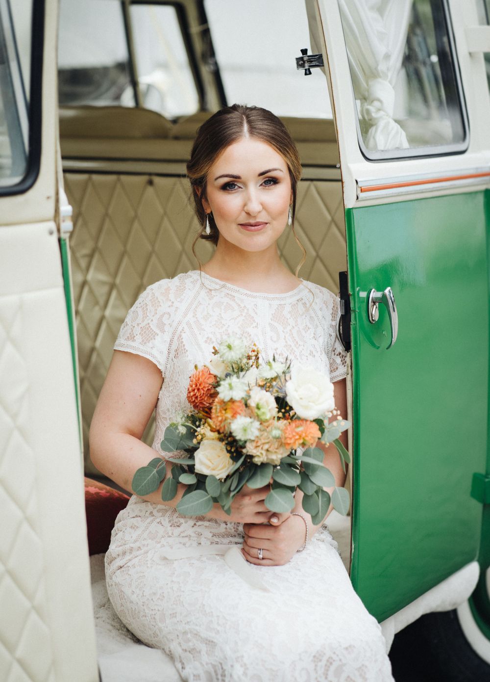 A person in a lace wedding dress holds a vibrant orange and white bouquet next to a vintage green and white van.