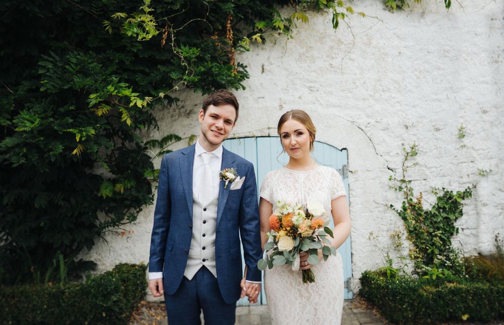 A wedding couple poses against a white wall with greenery, wearing formal attire and holding a colorful bouquet.