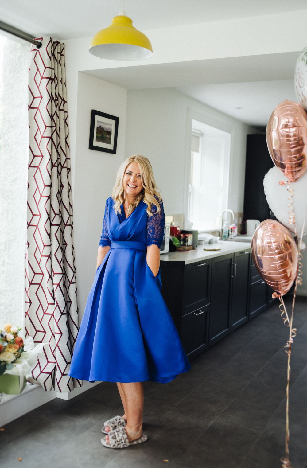 A woman in a royal blue midi dress stands in a modern kitchen with rose gold balloons and a yellow pendant light.