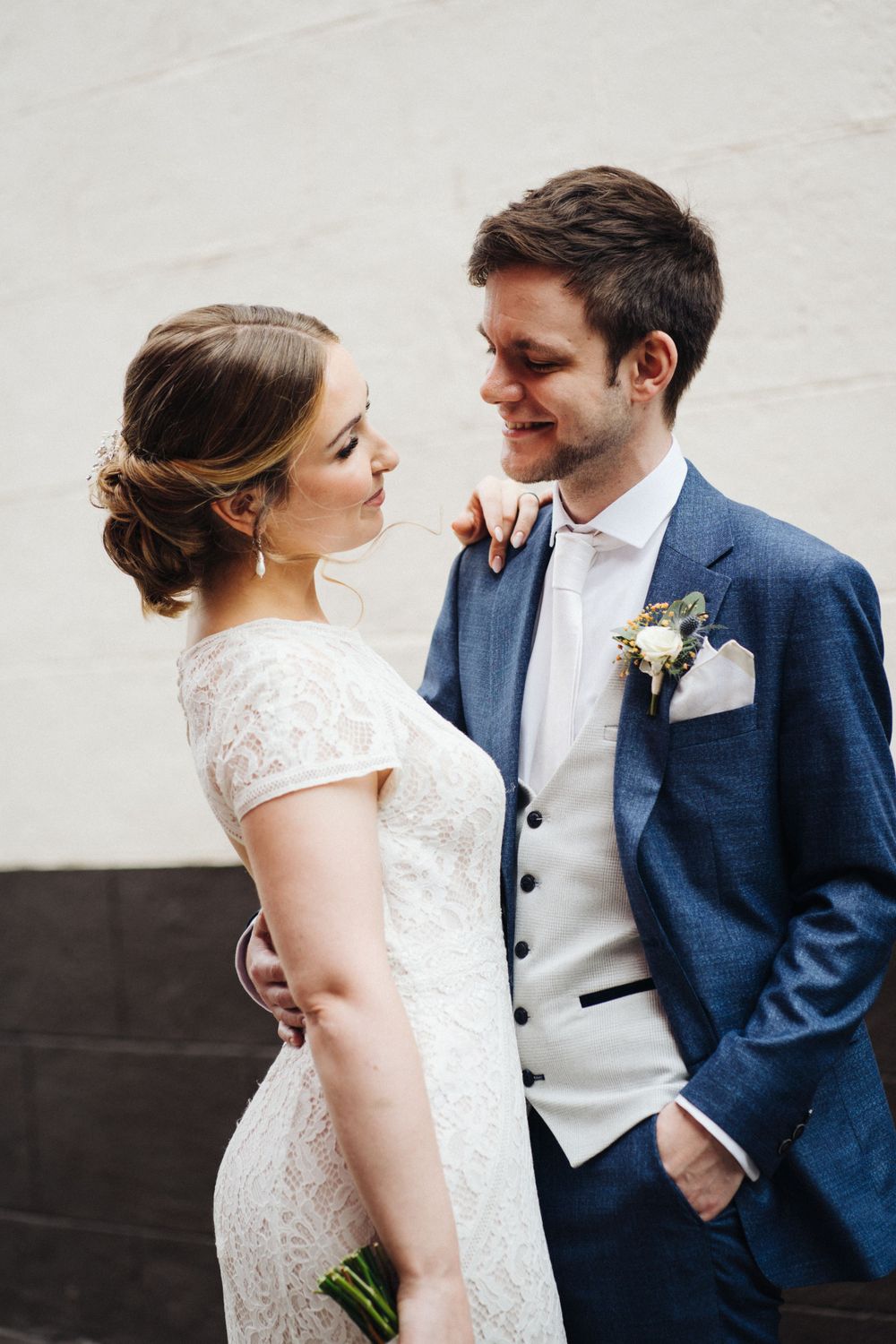 A romantic moment between newlyweds against a white wall, with the groom in navy and bride in lace.