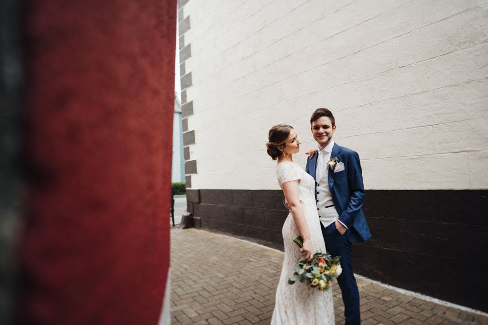 A couple in wedding attire stands against a geometric wall design holding a floral bouquet.